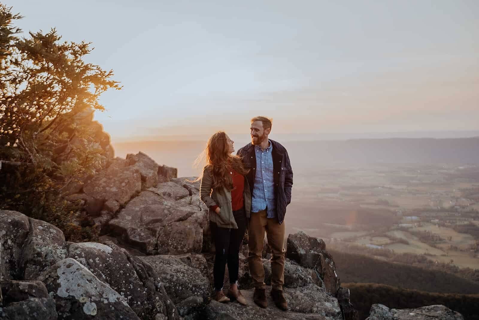 A couple stands on a huge rock in Shenandoah National Park while the sun sets behind them.