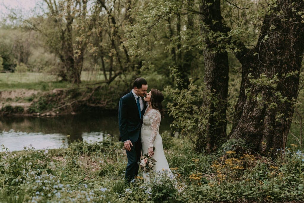 A newlywed couple stands in wildflowers along a riverbank