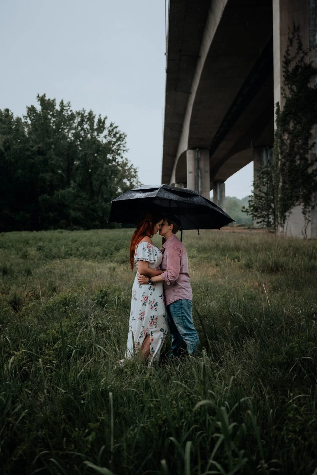 A lesbian couple kisses under a black umbrella in the middle of a field on belle isle in the middle of a rain storm.