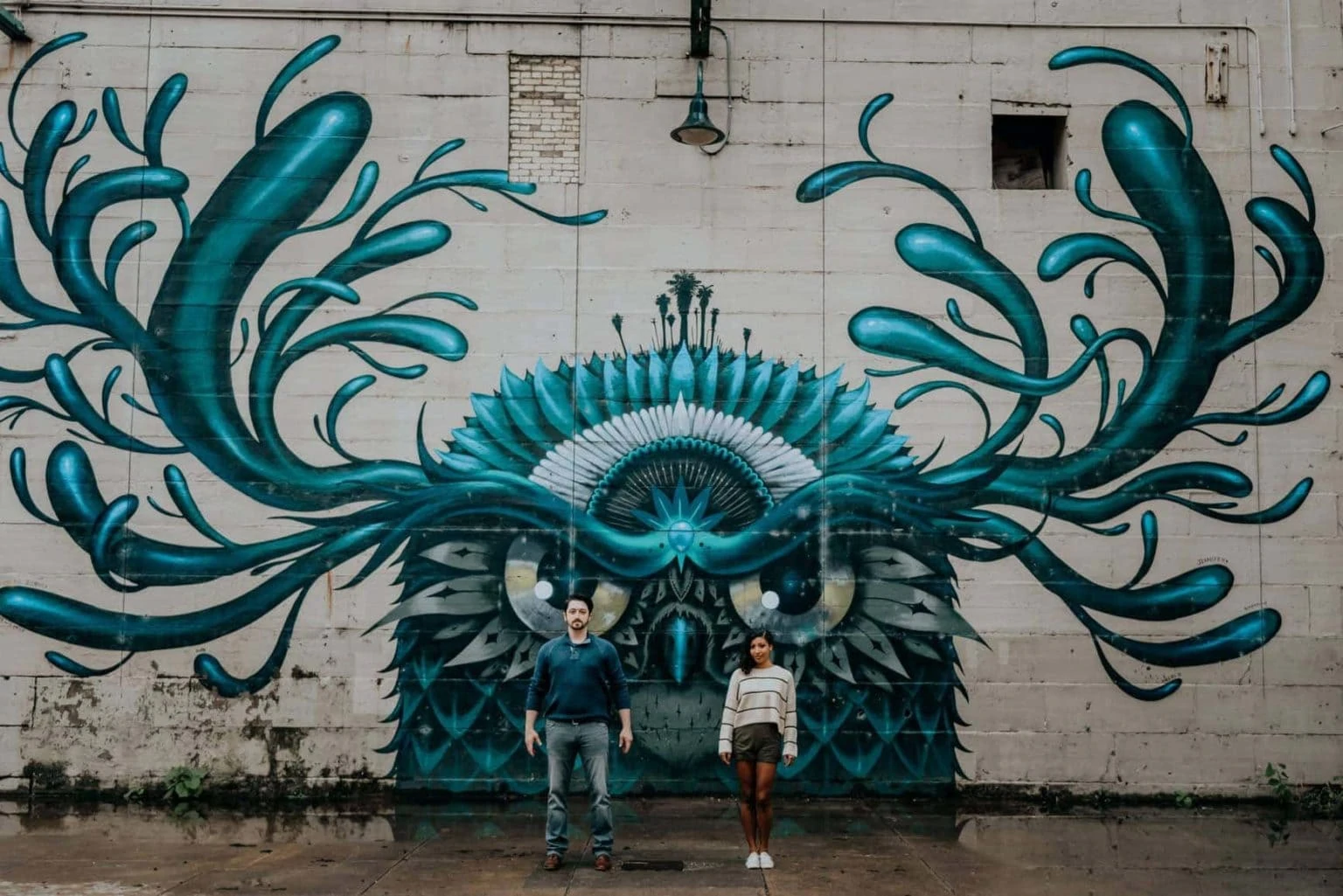 A couple stands in front of a mural of an owl-like creature