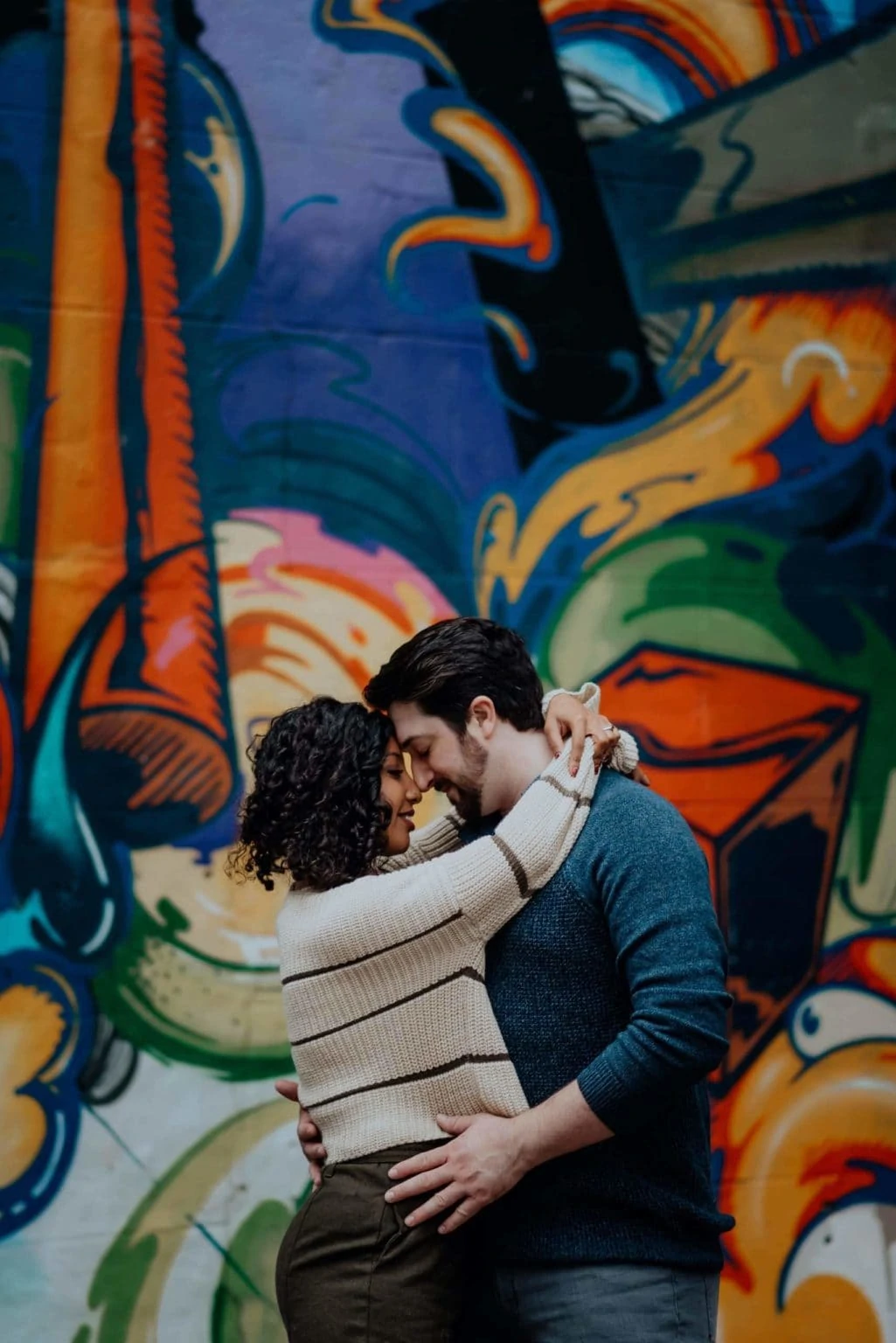 A couple embraces in front of a colorful mural.