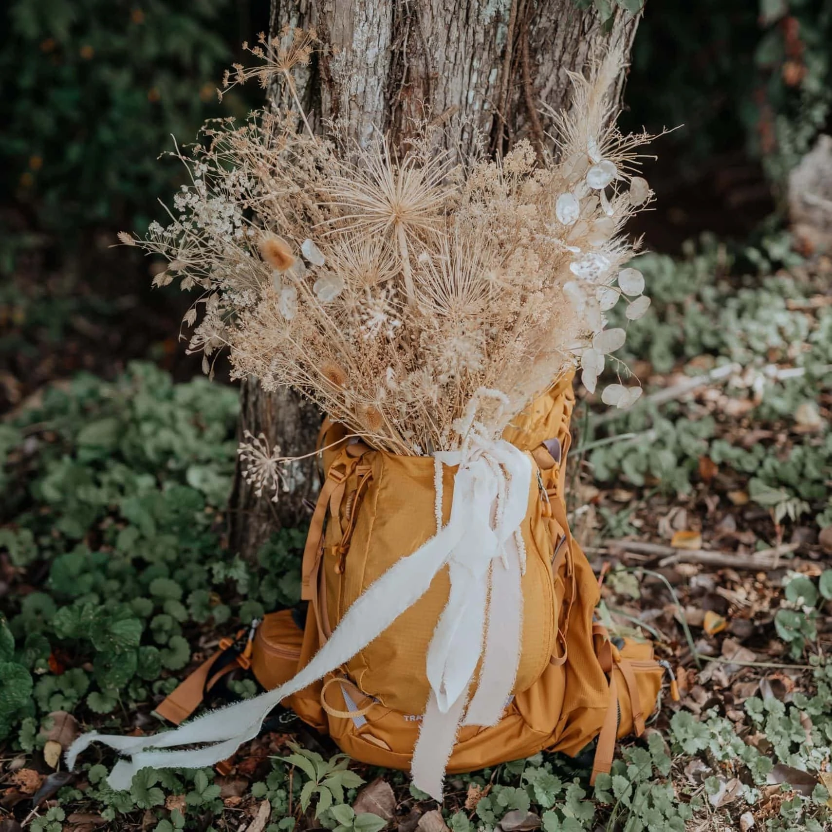 A bouquet of dried flowers and grasses is secured in a gold-colored hiking backpack for an elopement. Photographed by virginia elopement photographer, Flit Photography